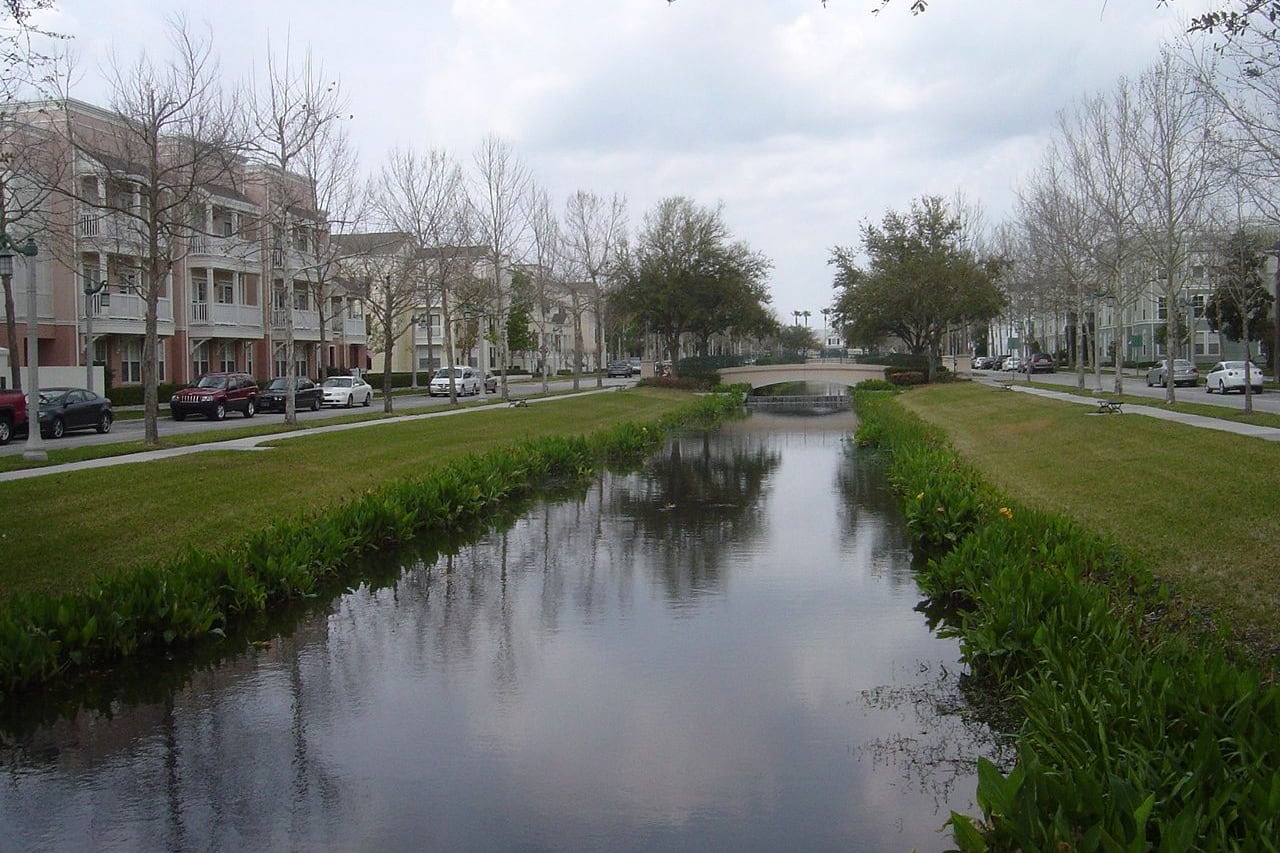 Canal com gramado dos dois lados, prédios residenciais e ponte de pedestre ao fundo