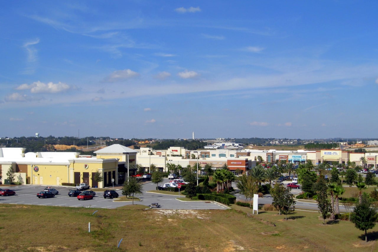 Centro comercial com lojas, estacionamento e palmeiras visto de longe.