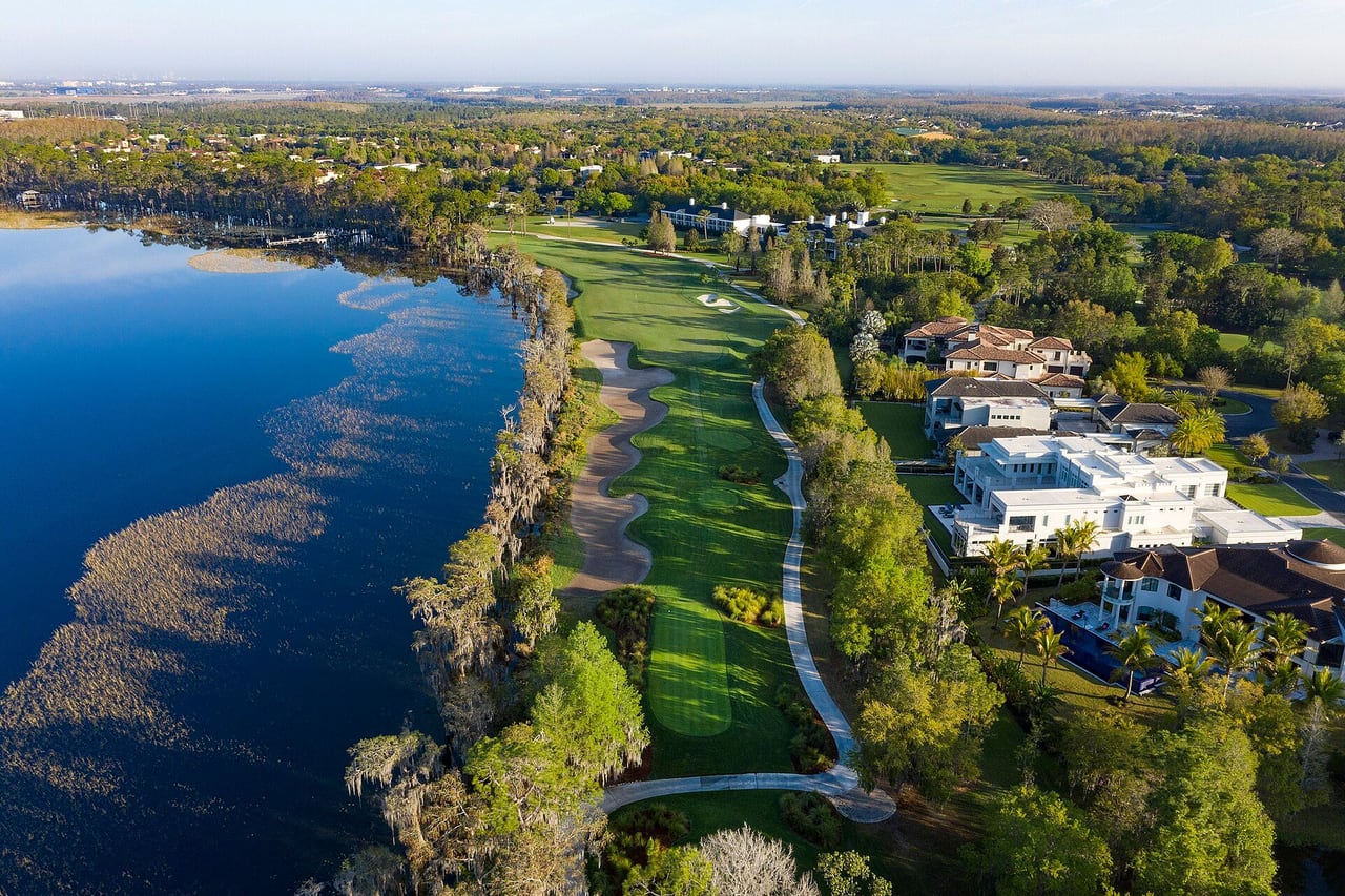 Vista do alto de um campo de golfe entre um lago e casas grandes cercadas de árvores