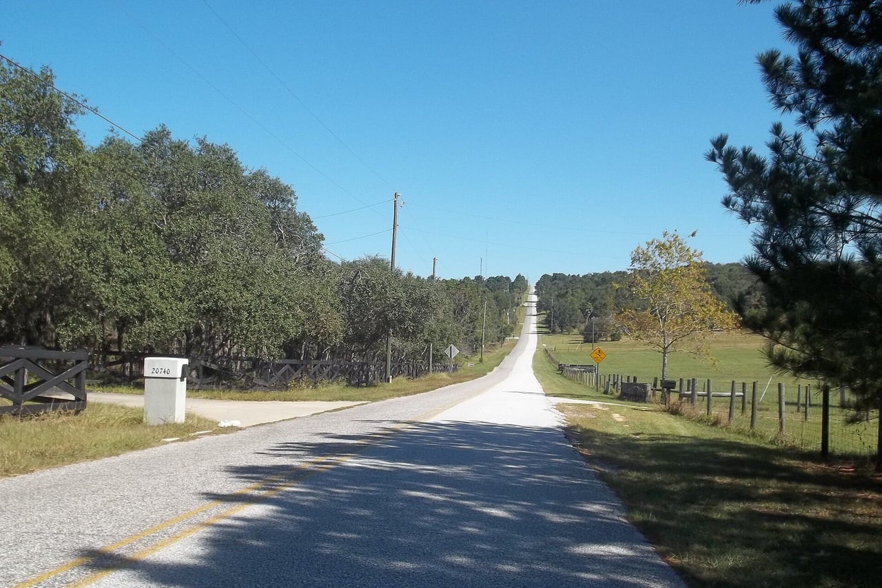 Estrada rural com cerca de madeira, caixa de correio e pasto ao lado.